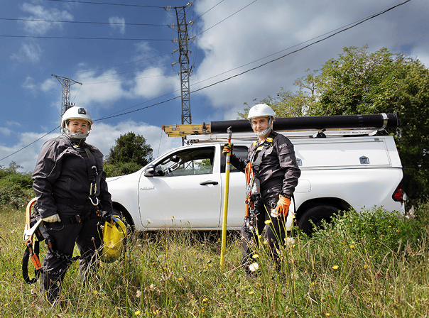 two utility workers wearing safety gear, including helmets and gloves, stand in front of a white vehicle equipped with mounted tools in a grassy area. behind them are power lines and a transmission tower, suggesting maintenance or repair operations in an electrical field