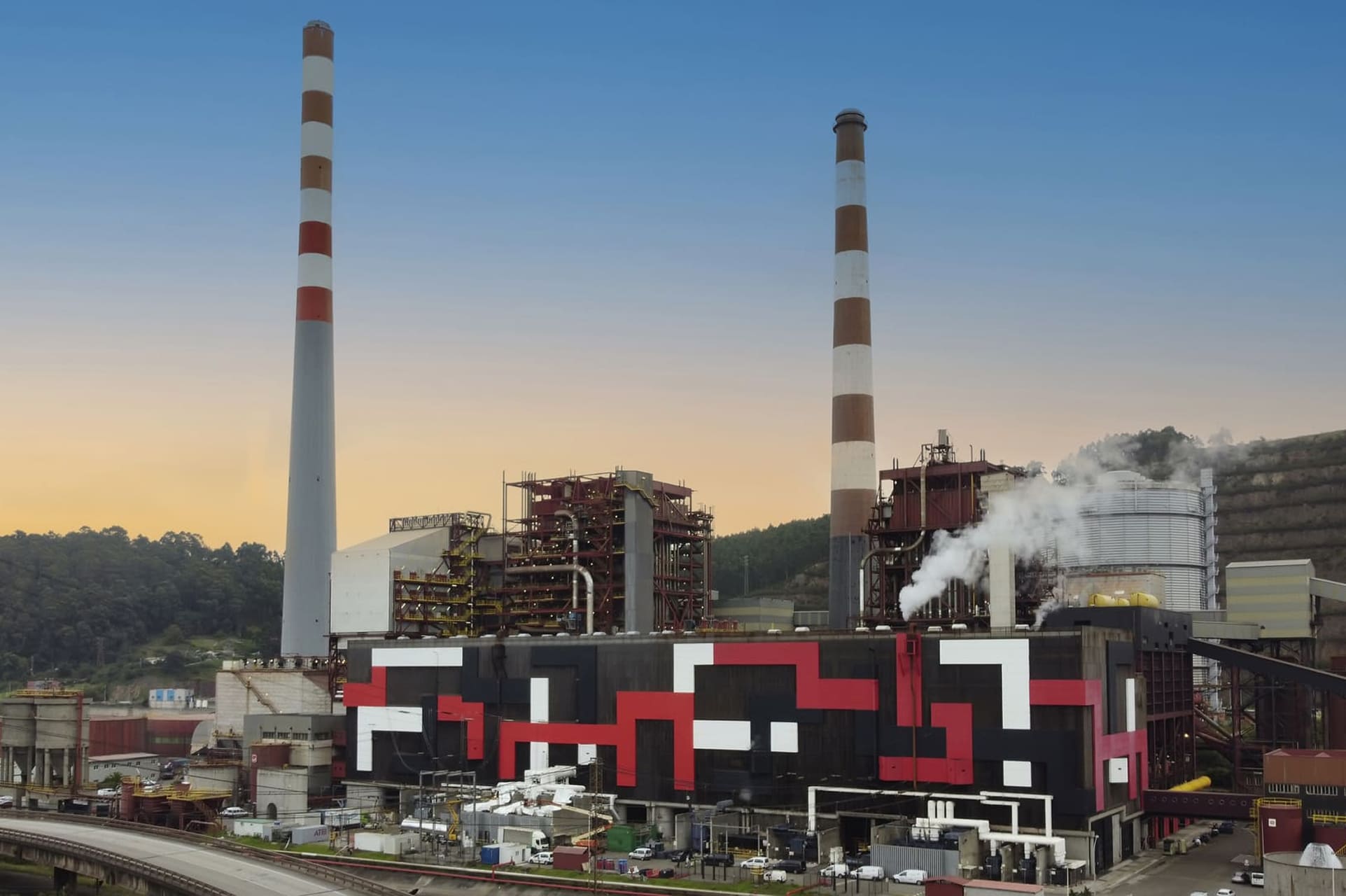 industrial site of a thermal power plant with two tall chimneys and geometric patterns on the exterior, surrounded by greenery and a clear sky