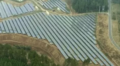 a large solar farm with rows of solar panels arranged in a grid pattern on sloped terrain. the setup is surrounded by trees, and a road runs along the right side of the image, connecting the infrastructure to its surroundings