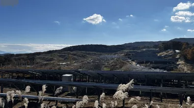 a solar farm located on sloped terrain with rows of panels positioned to maximize sunlight absorption. hills covered with trees and vegetation are visible in the background, under a bright, partly cloudy sky