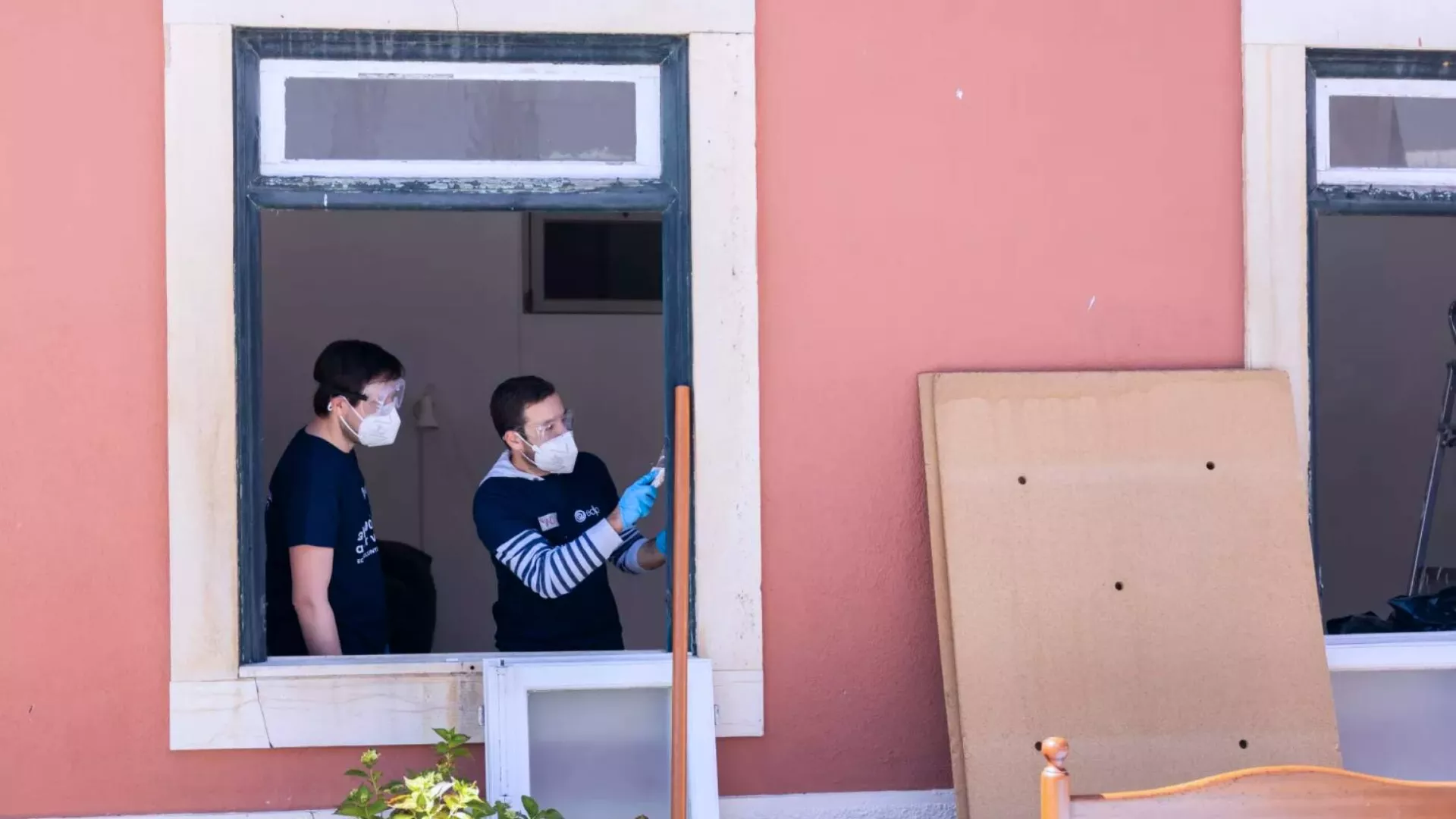 two volunteers working on a project, seen through a window. one person is painting a window frame with a brush, wearing gloves and a mask, while another observes or assists. outside, a piece of cardboard leans against a wall, suggesting renovation or cleanup efforts