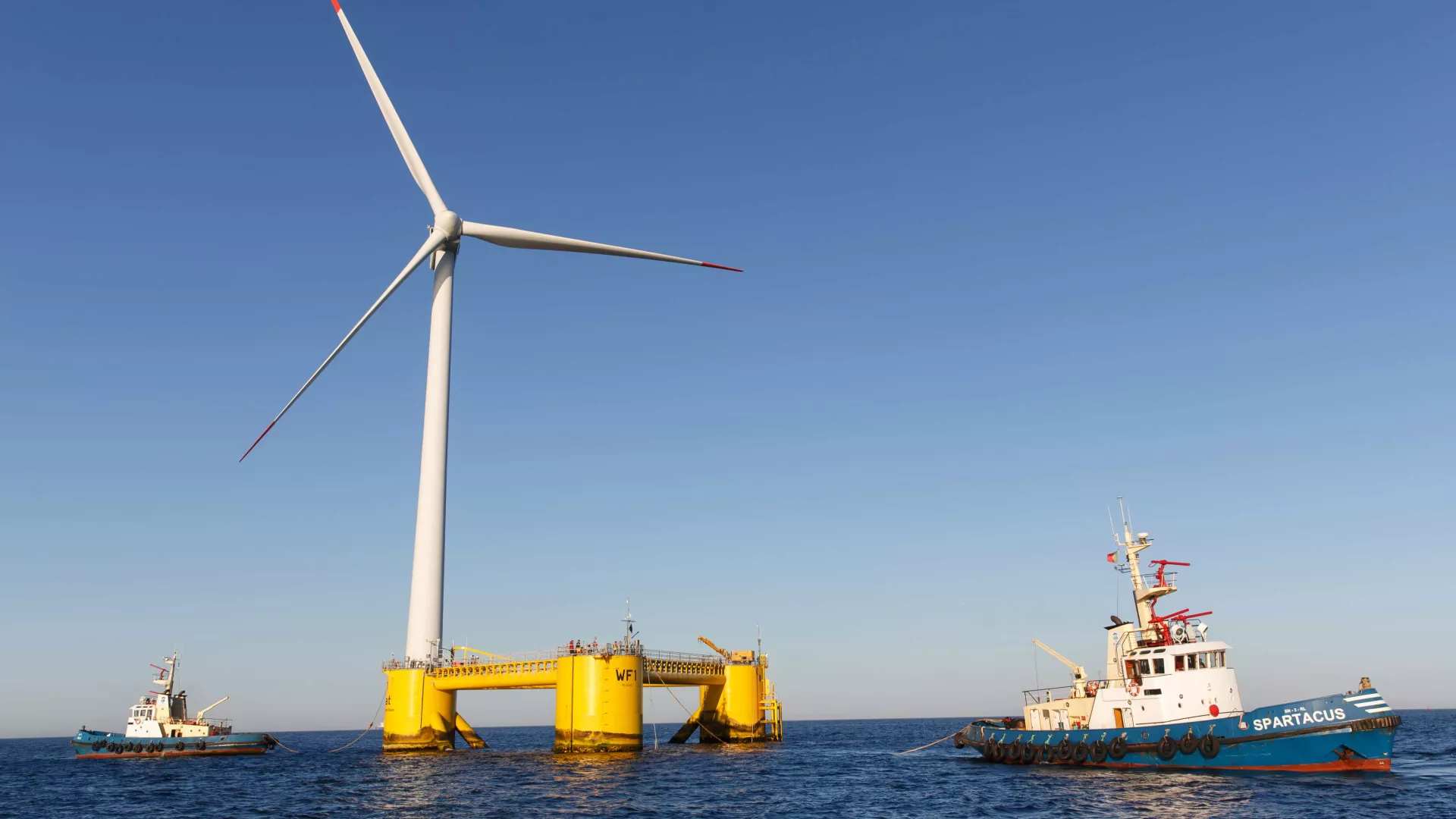 floating wind turbine mounted on a yellow platform labeled 'efi,' anchored in calm ocean waters with a boat nearby